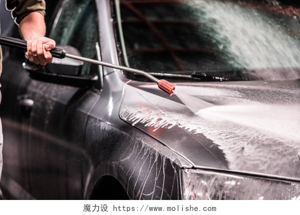 男人拿着机器在冲洗轿车一个人用胡子或洗车机洗一辆灰色的汽车与高压垫圈在晚上在商店洗涤